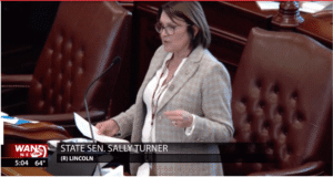 State Senator Sally Turner speaks at a podium on the Senate floor introducing a Celiac Disease Training Bill. She is wearing glasses and a plaid blazer, holding papers while addressing the chamber. The screen graphic reads “STATE SEN. SALLY TURNER (R) LINCOLN” with a WAND News logo in the bottom left corner showing the time and temperature. 